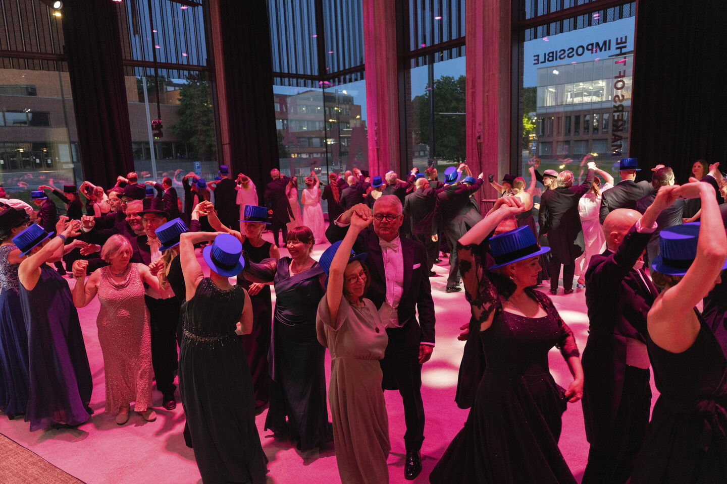 A formal event with people dancing. Many are wearing blue hats, dark evening gowns, and suits. Tall windows show an urban view.
