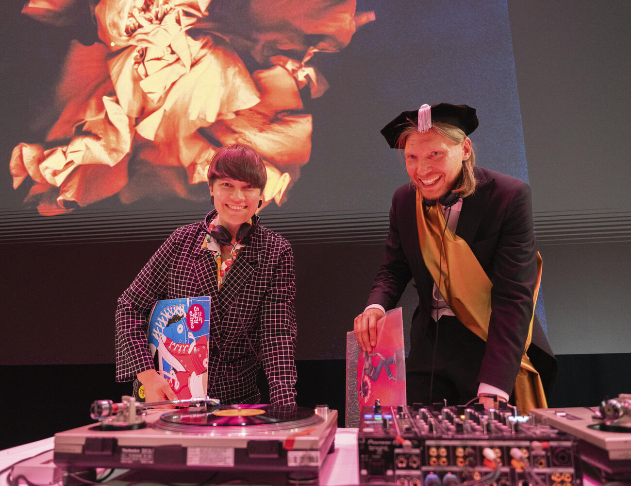 Two people next to DJ equipment, one in a checkered outfit, the other in academic dress. Abstract art is projected behind them.