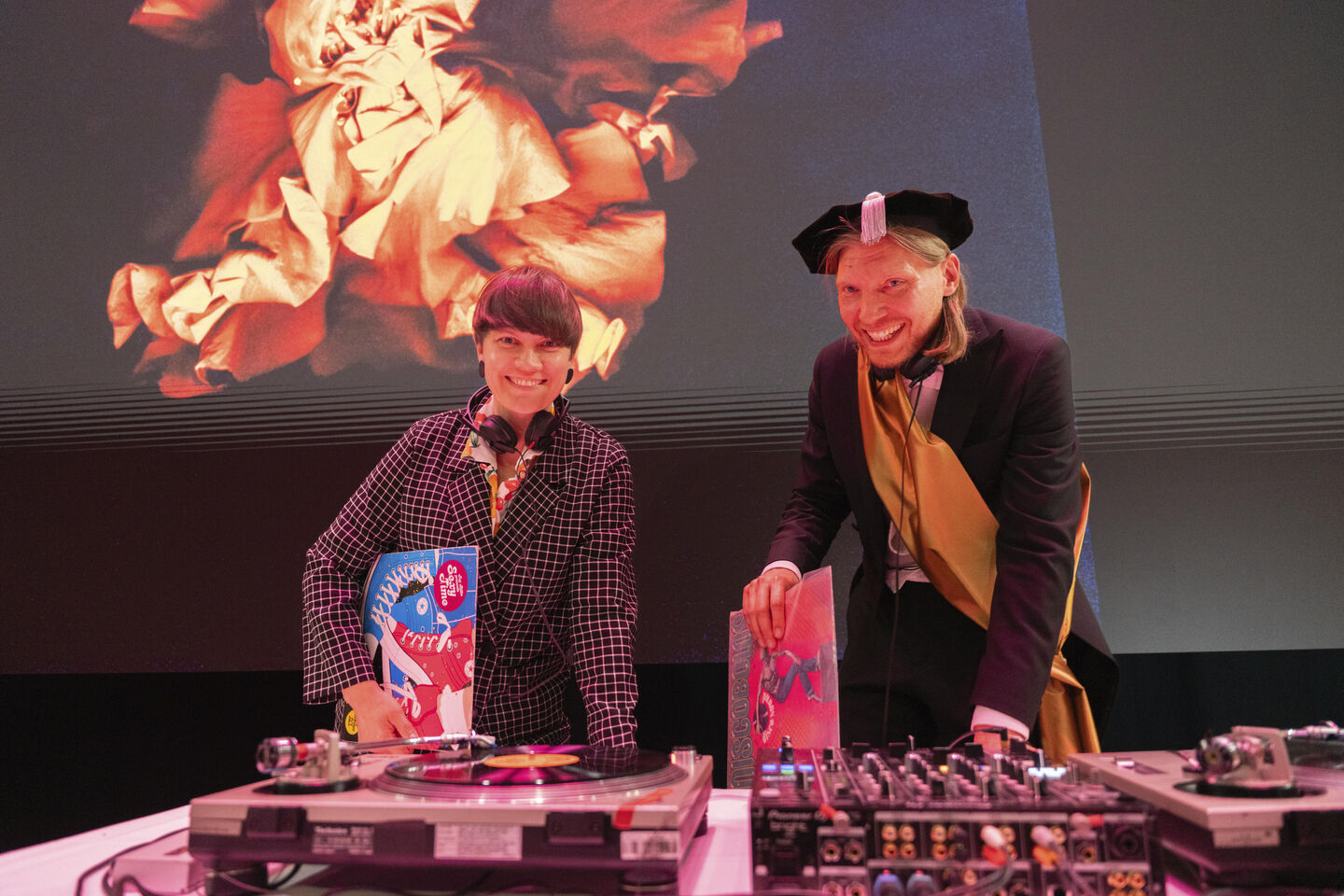 Two people next to DJ equipment, one in a checkered outfit, the other in academic dress. Abstract art is projected behind them.
