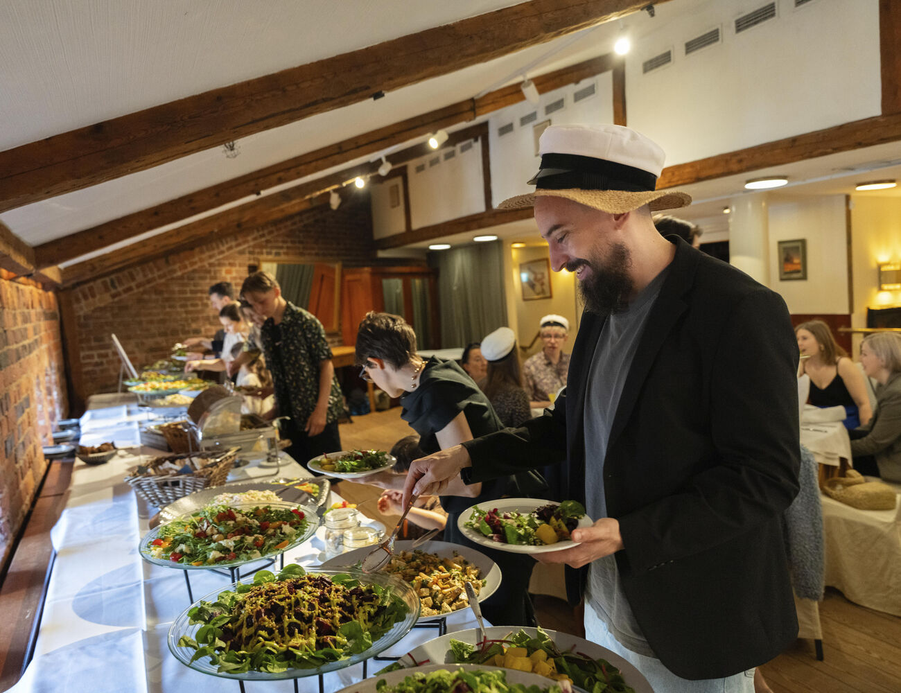 People in casual attire enjoy an indoor buffet. The room has exposed beams and brick walls.