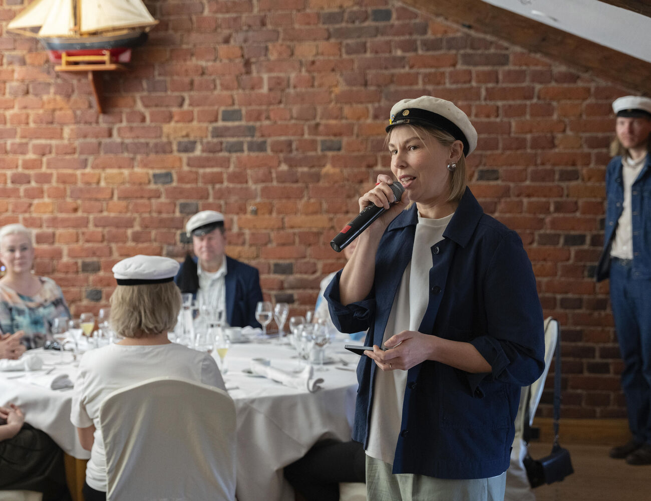 Person dressed in casual outfit with white hat speaking into a microphone. Attendees are seated at a round table in front.