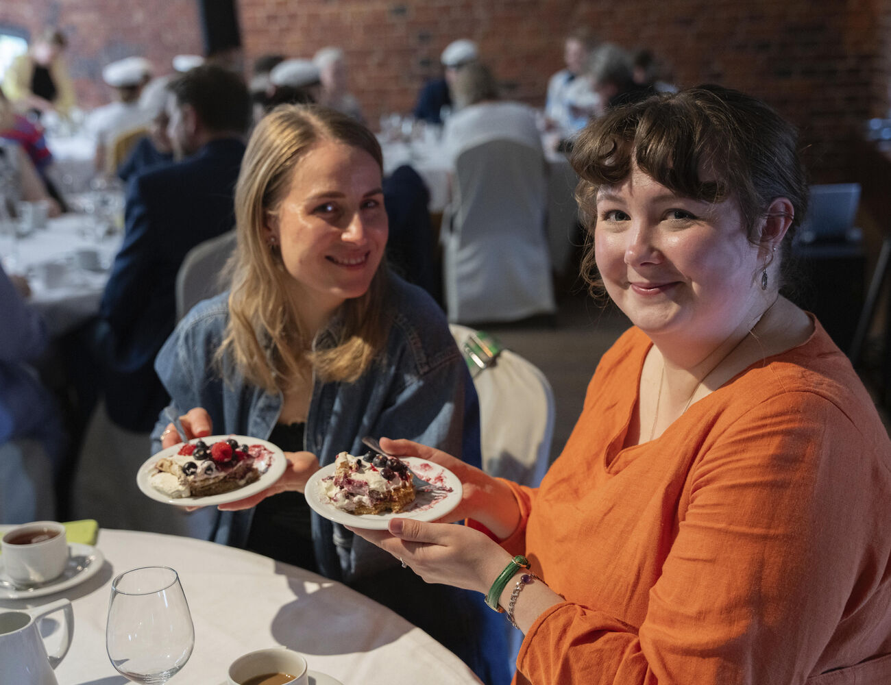 Two people holding dessert plates at a gathering. One wears an orange shirt, the other a denim jacket. Table with cups and glasses.