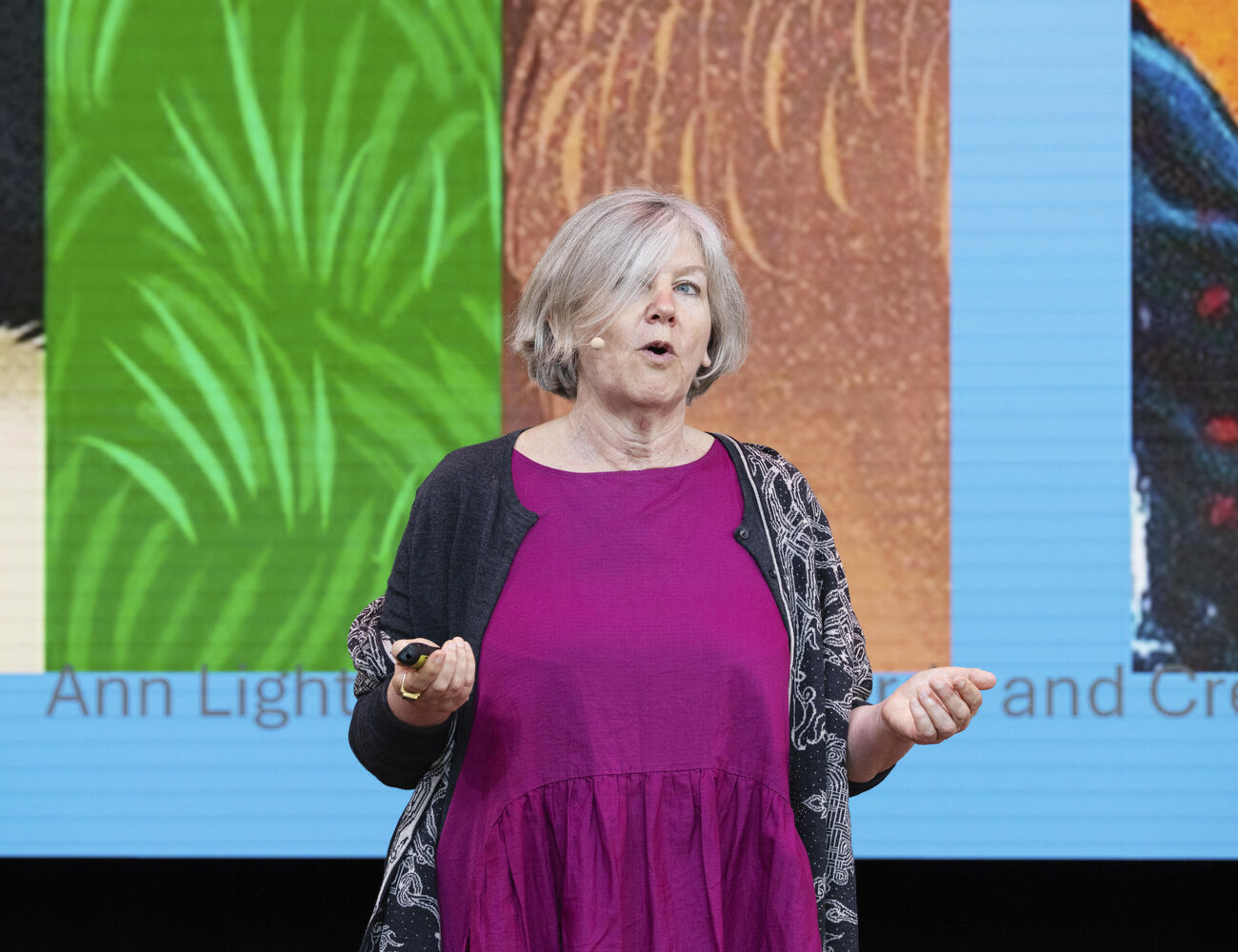 A woman in a purple dress and dark cardigan stands in front of a colourful background screen.