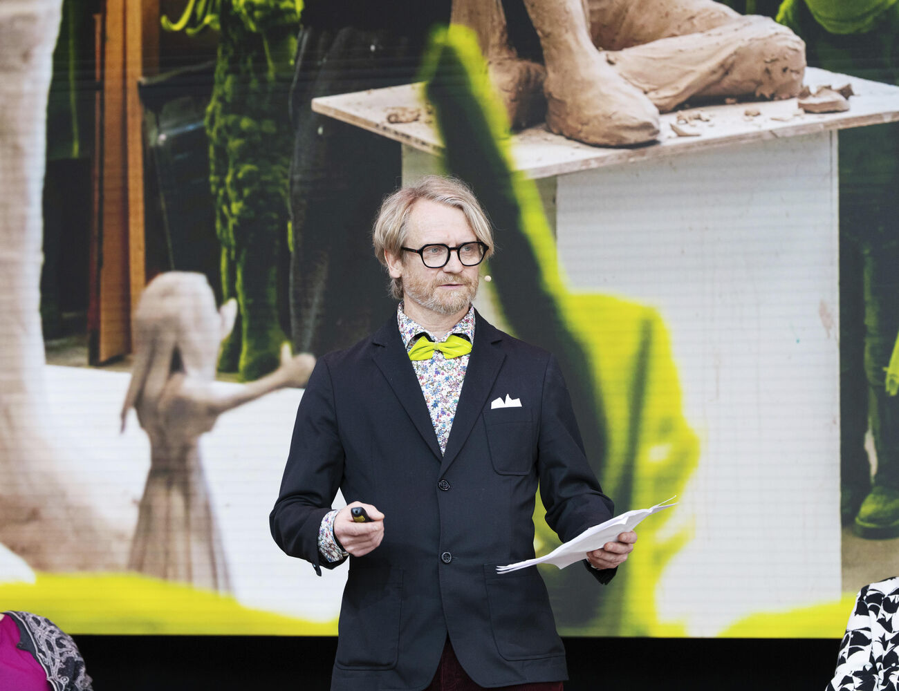 Person in a suit holding papers speaks on stage with an art sculpture and a painting in the background.
