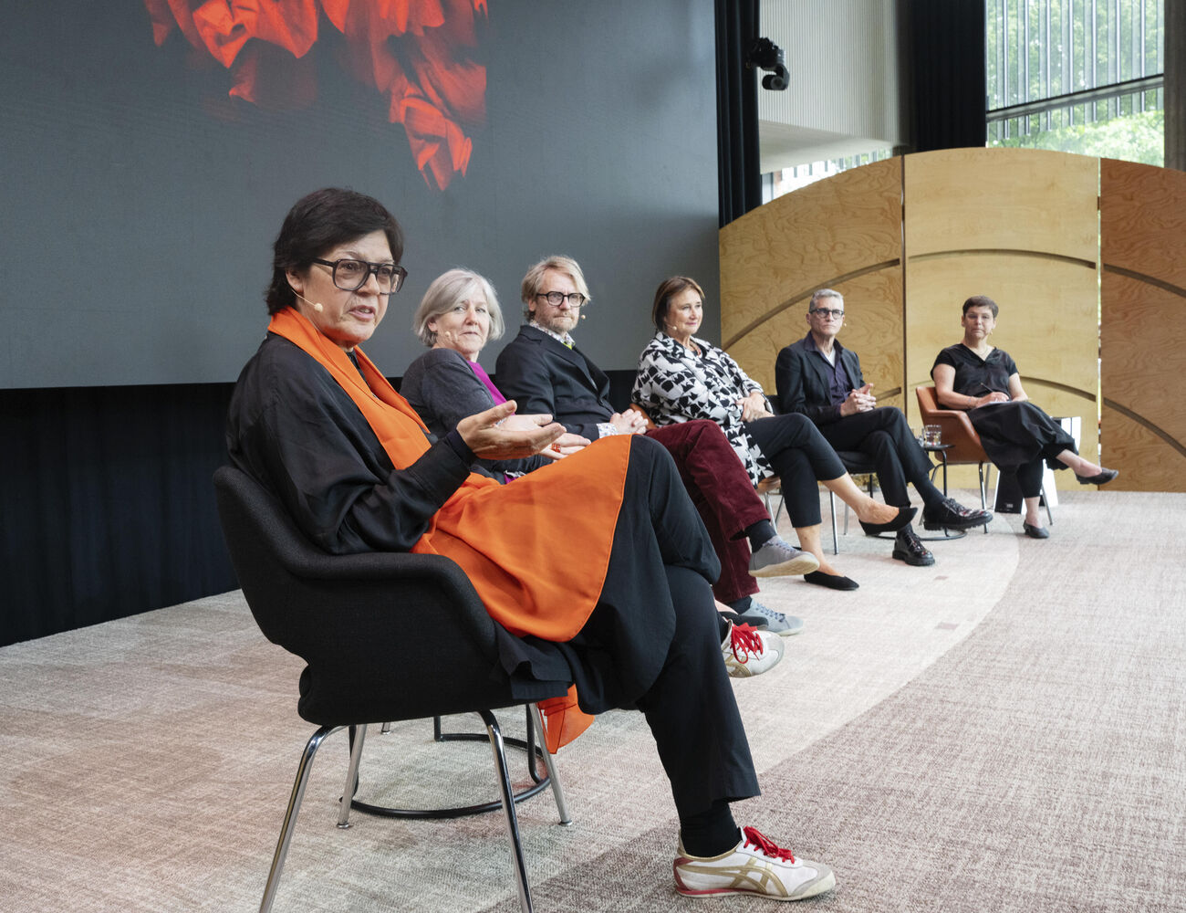 Six people seated in a panel discussion. The person in the foreground wears a black outfit with an orange scarf.