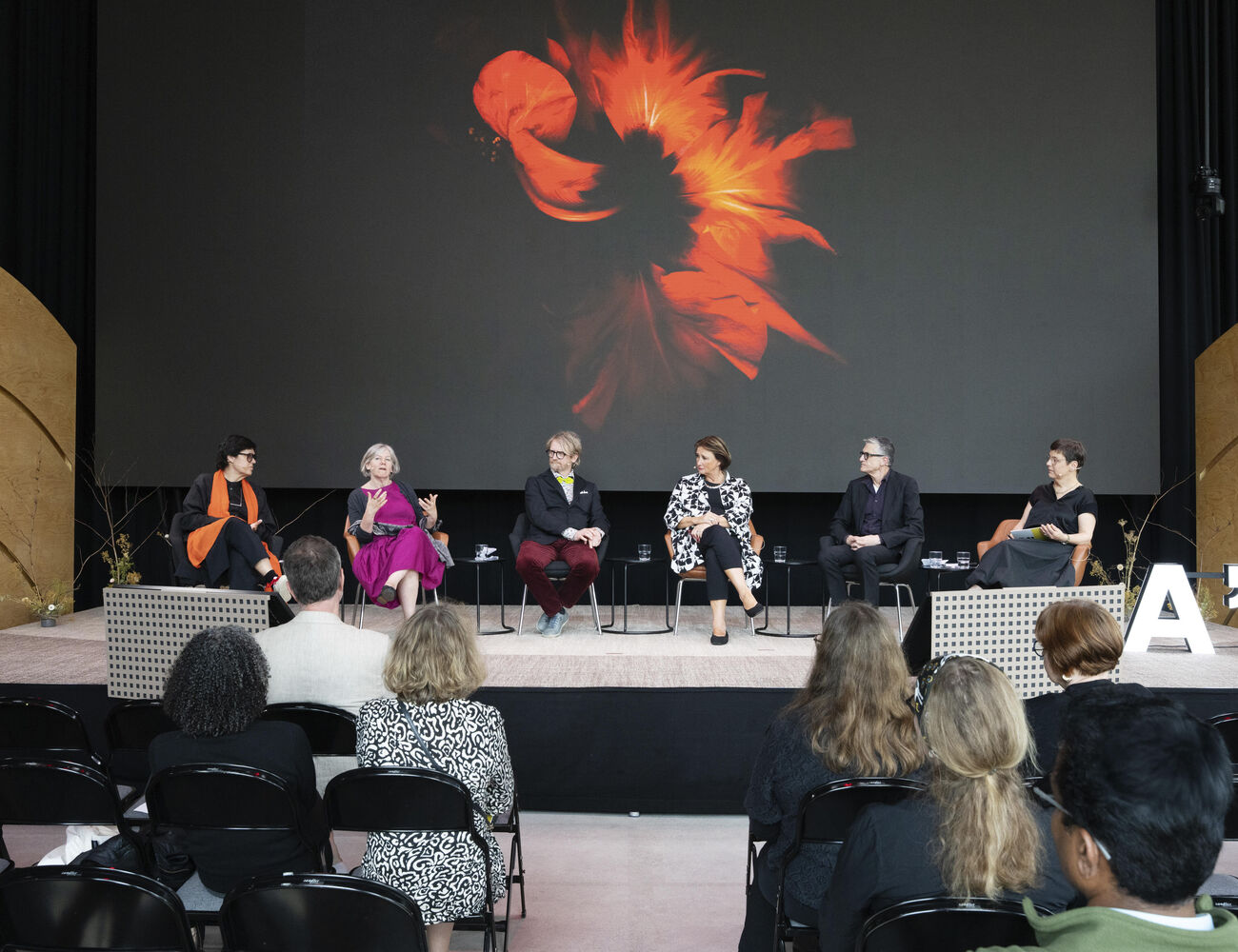 Six people sit on stage discussing, behind them a large screen showing abstract floral art. Audience watches attentively.