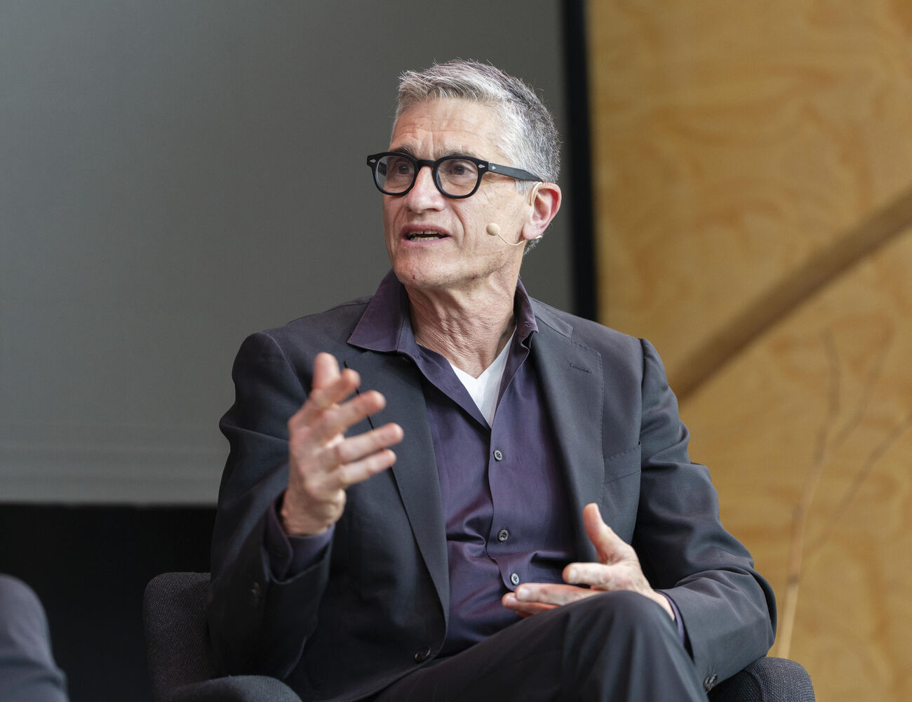 Person in a suit gesturing with hand while seated indoors.