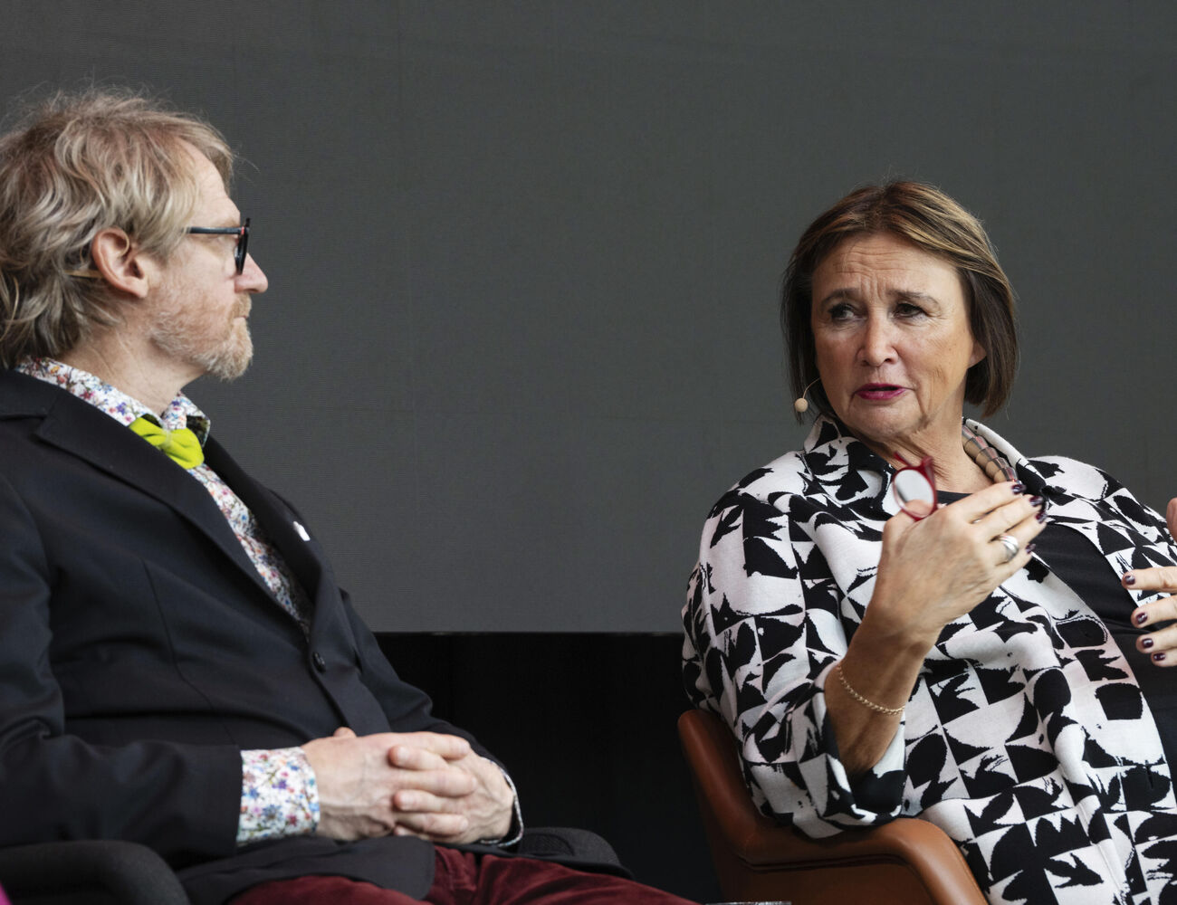 Two people seated in armchairs, one wearing a black blazer with a green bow tie, the other in monochrome patterned clothing.