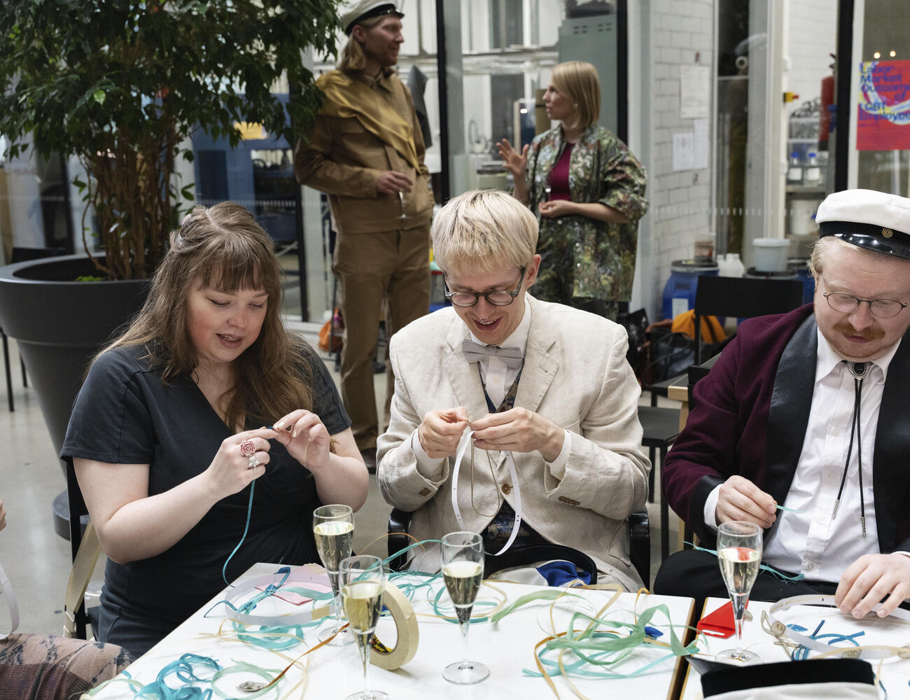 Three people seated at a table, crafting with ribbons and drinking champagne. Two people standing in the background.