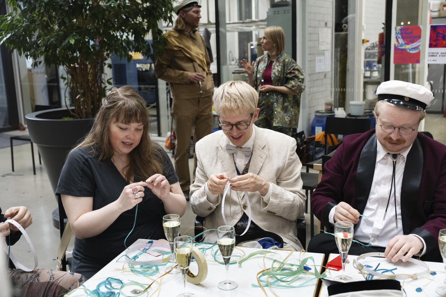 Three people seated at a table, crafting with ribbons and drinking champagne. Two people standing in the background.