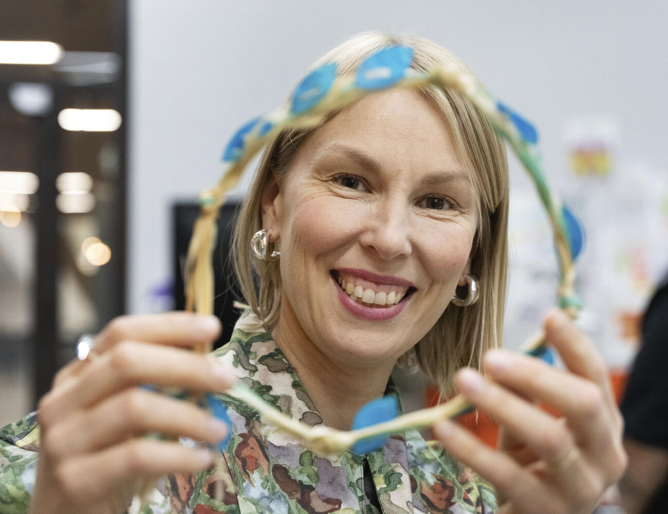 Person holding a handmade wreath decorated with blue petals, wearing a camouflage-patterned shirt indoors.