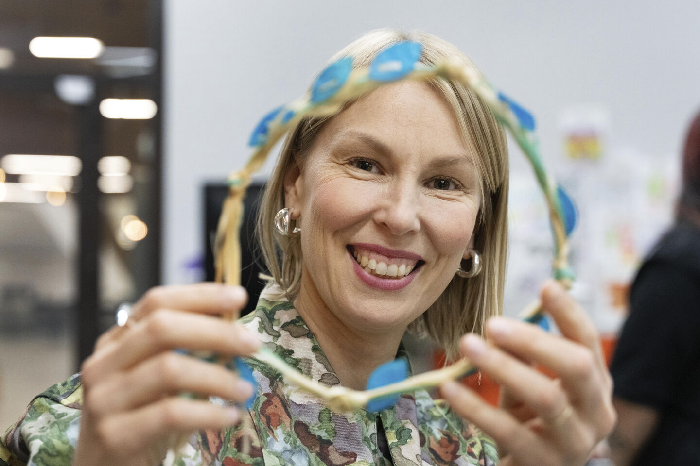 Person holding a handmade wreath decorated with blue petals, wearing a camouflage-patterned shirt indoors.