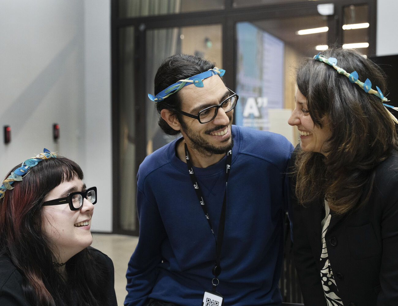 Three people indoors wearing blue flower crowns and dark clothing, with lanyards around their necks.
