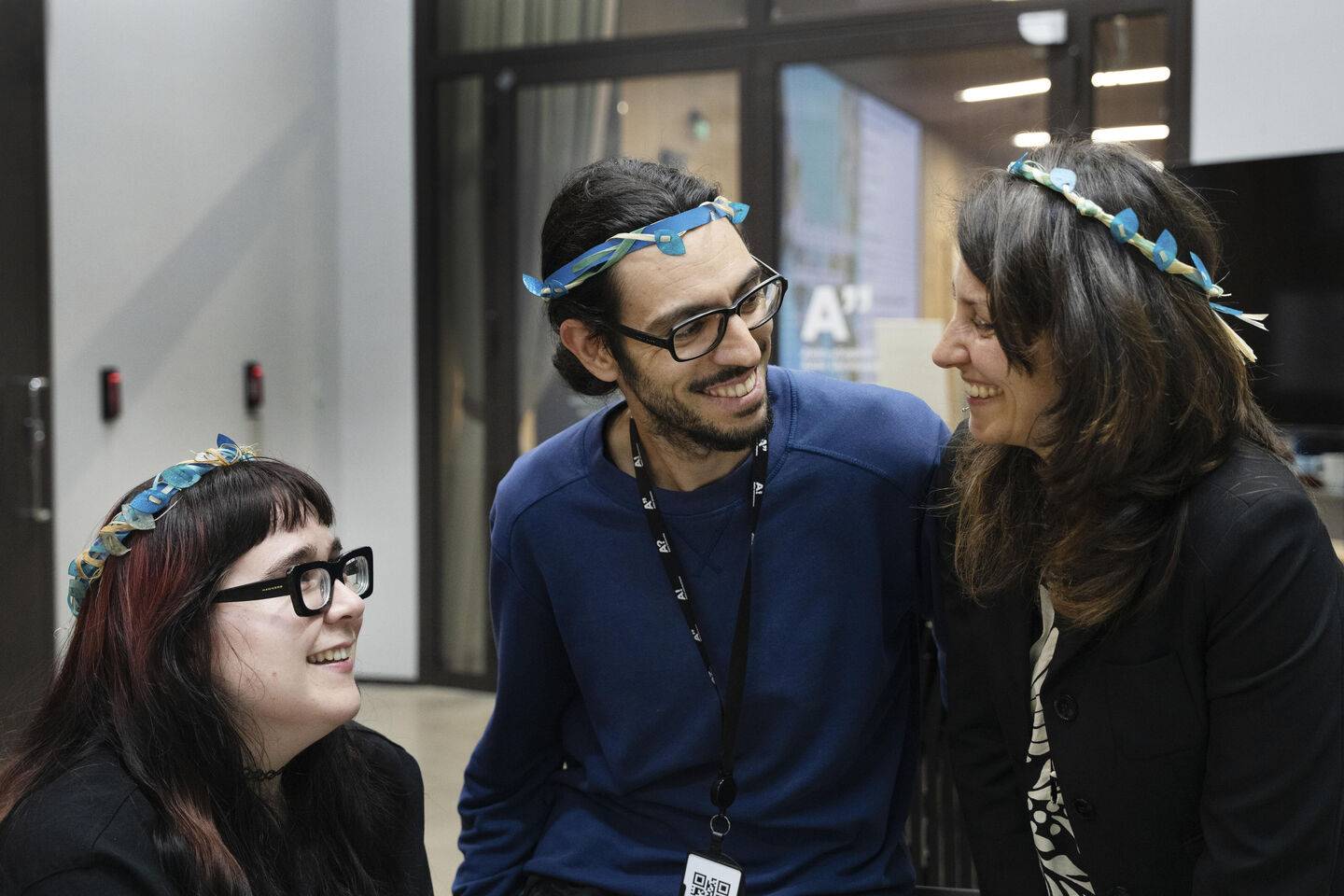 Three people indoors wearing blue flower crowns and dark clothing, with lanyards around their necks.