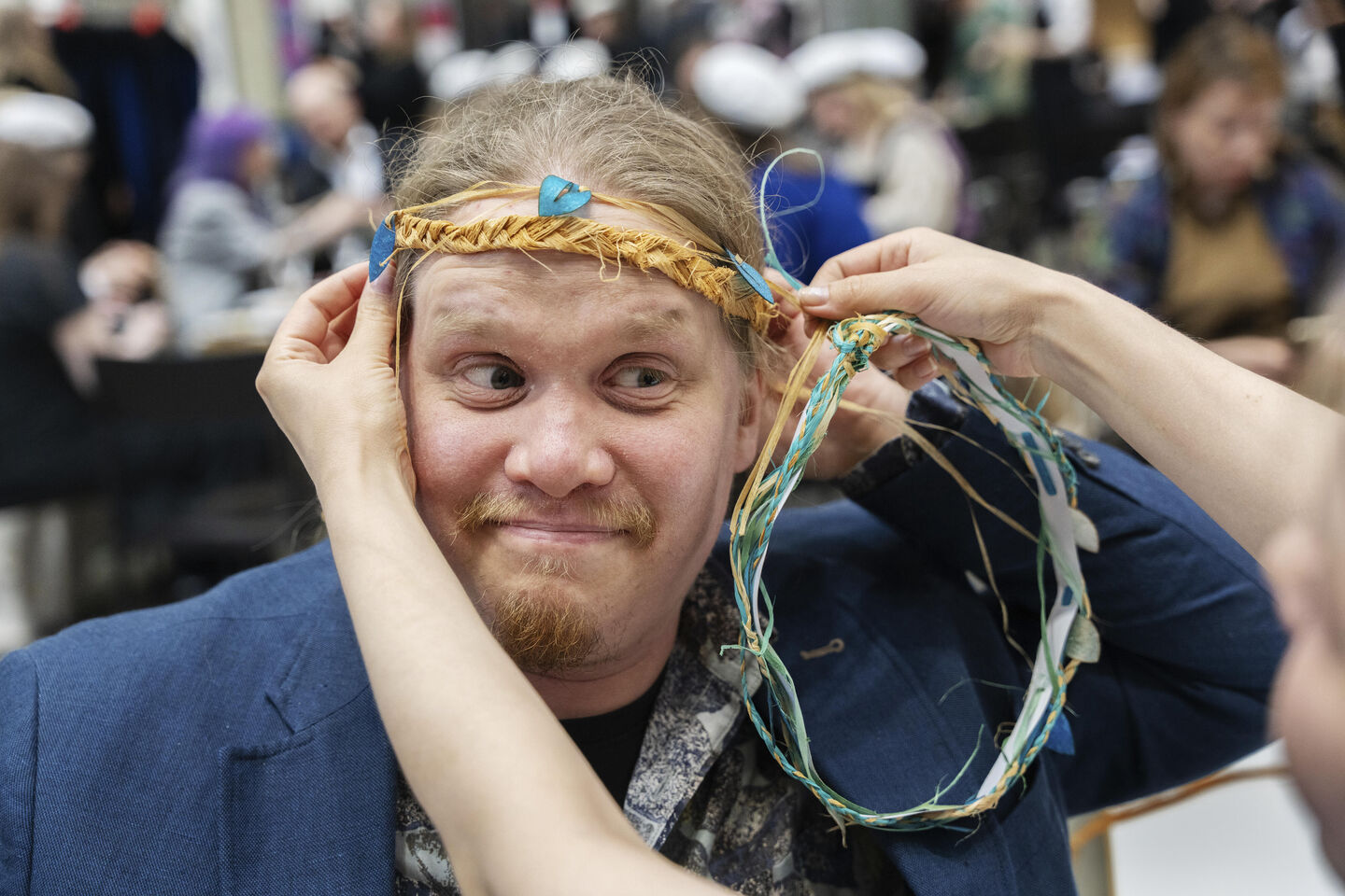 A person wearing a blue jacket is being crowned with a wreath made of colourful ribbons at a crowded indoor event.