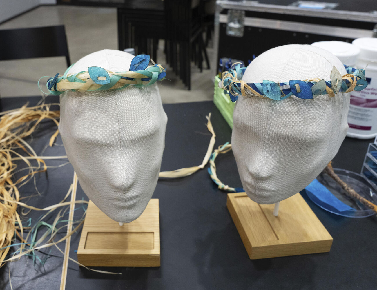 Two head mannequins with handmade wreaths of dried plants and blue paper leaves on a table.