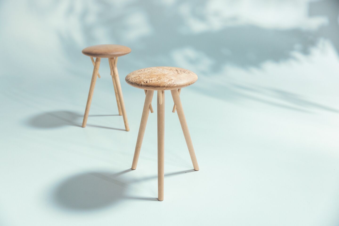 Two wooden stools with three legs each, standing on a light blue surface with shadows.