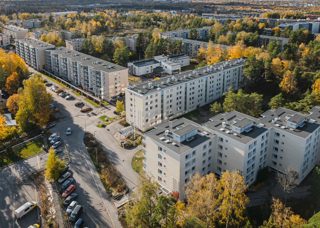 Ilmakuva kerrostaloista kaupunkialueella, jota ympäröivät puut ja syksyn lehdet. Katujen varrella on pysäköityjä autoja.