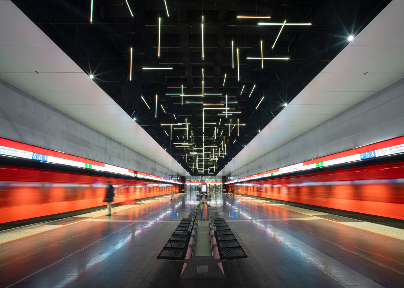 Moderni metroasema, jossa on punaiset junat ja taiteelliset kattovalot. Henkilö kävelee asemalaiturilla.