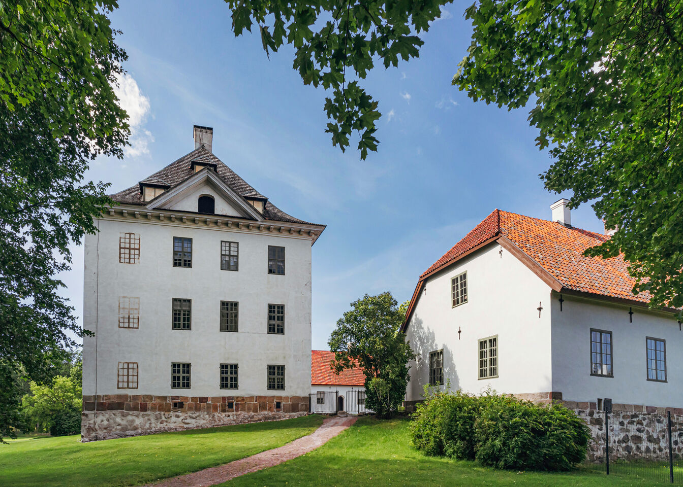 Valkoinen kivirakennus, jolla korkea katto, ja viereinen talo punatiilikatolla nurmialueella, jossa puita.