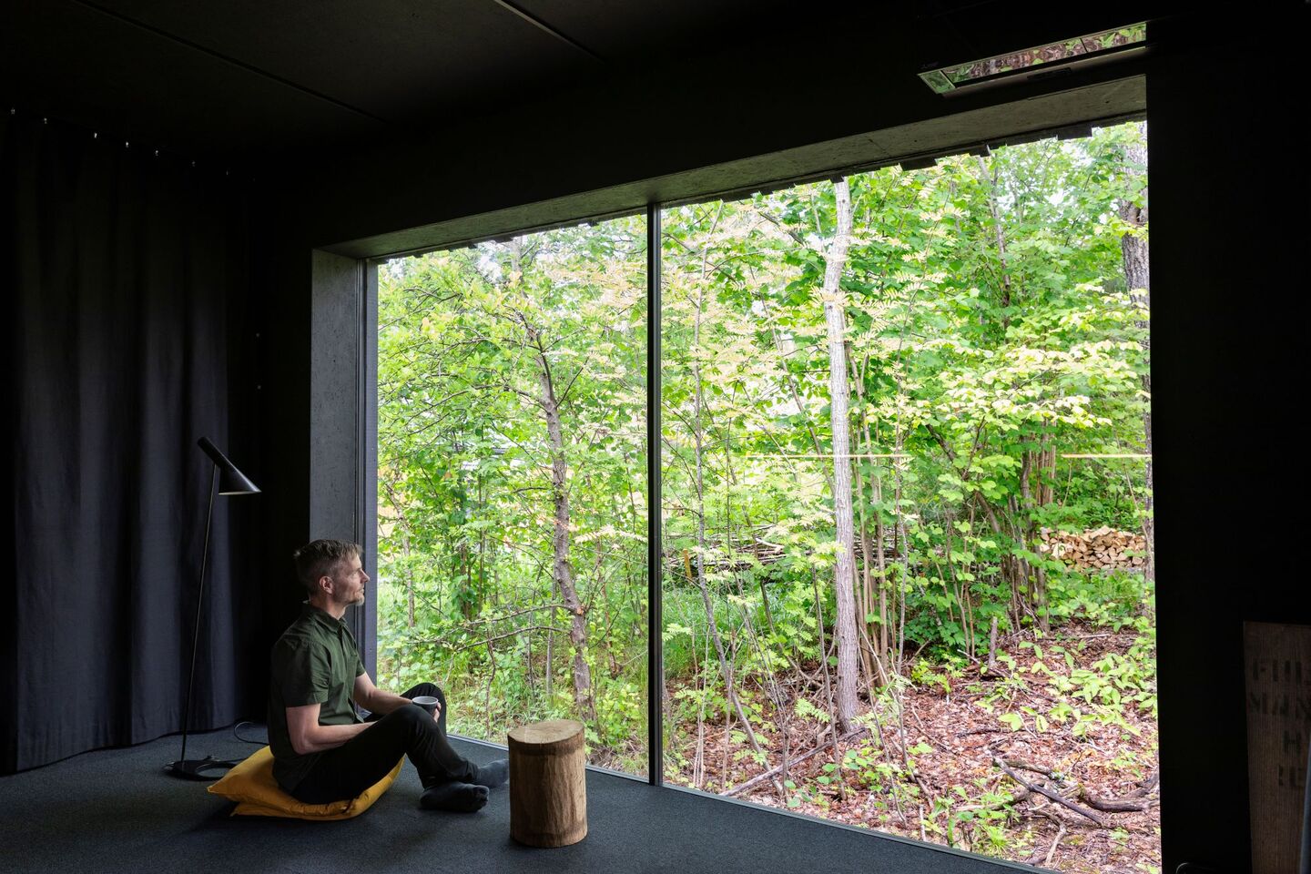 Person sitting indoors with large glass windows overlooking a lush green forest, next to a floor lamp and a wooden stump.
