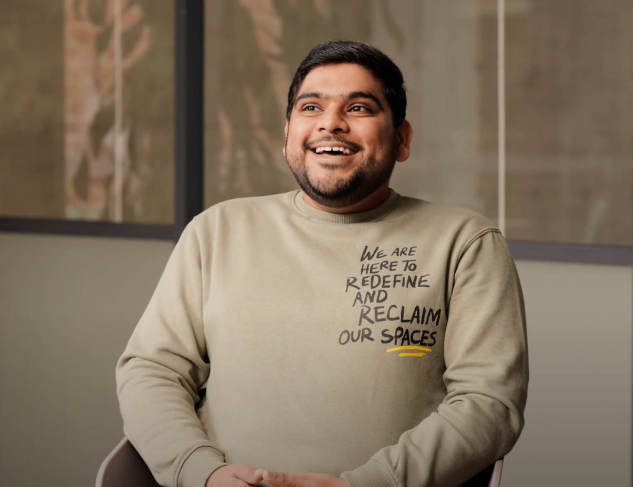 Person sitting indoors with a smile on their face, wearing a beige sweatshirt with text: 'We are here to redefine and reclaim our spaces.'