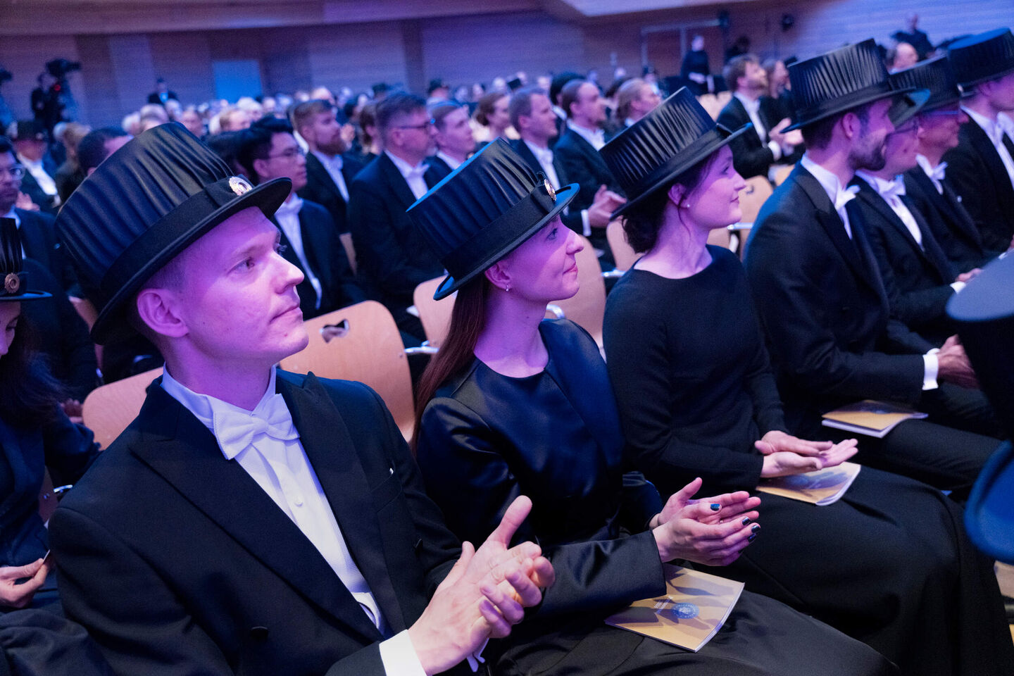 People wearing academic caps and formal attire seated at an indoor event.