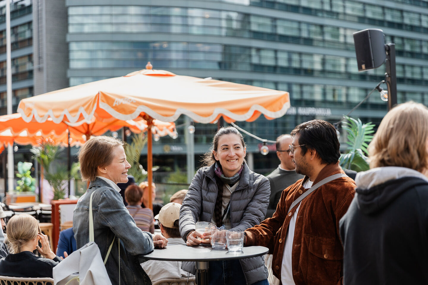 People socialising at an outdoor cafe with orange umbrellas and a modern building in the background.