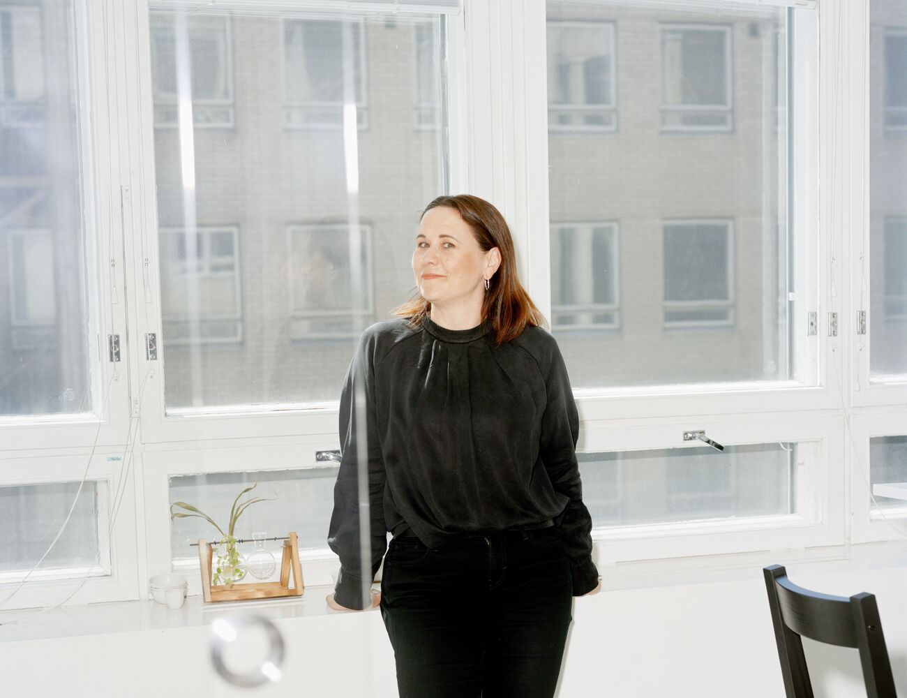A person stands indoors by a white counter in front of tall windows. The person is dressed in black clothing and shoes.
