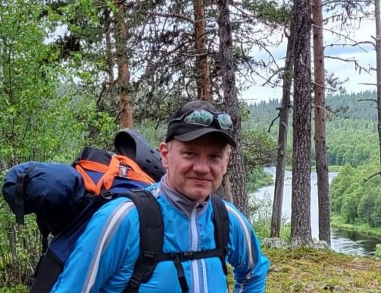 Siniseen takkiin ja lippalakkiin pukeutunut henkilö vaeltaa ison repun kanssa. Taustalla näkyy puita ja joki.