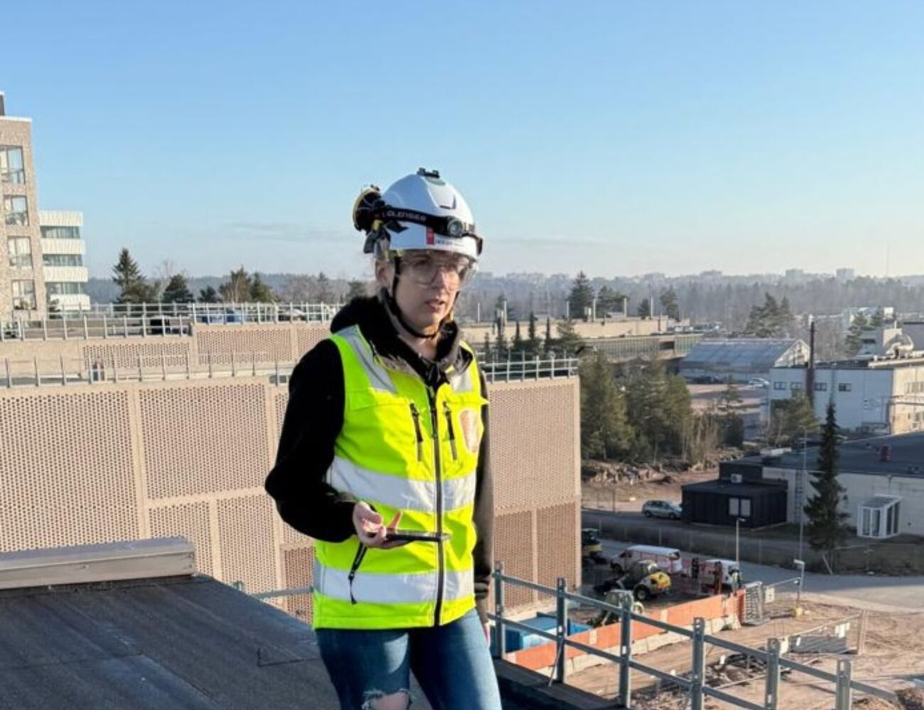 Person wearing high-visibility vest and hard hat, standing on a rooftop near construction site. Blue sky.