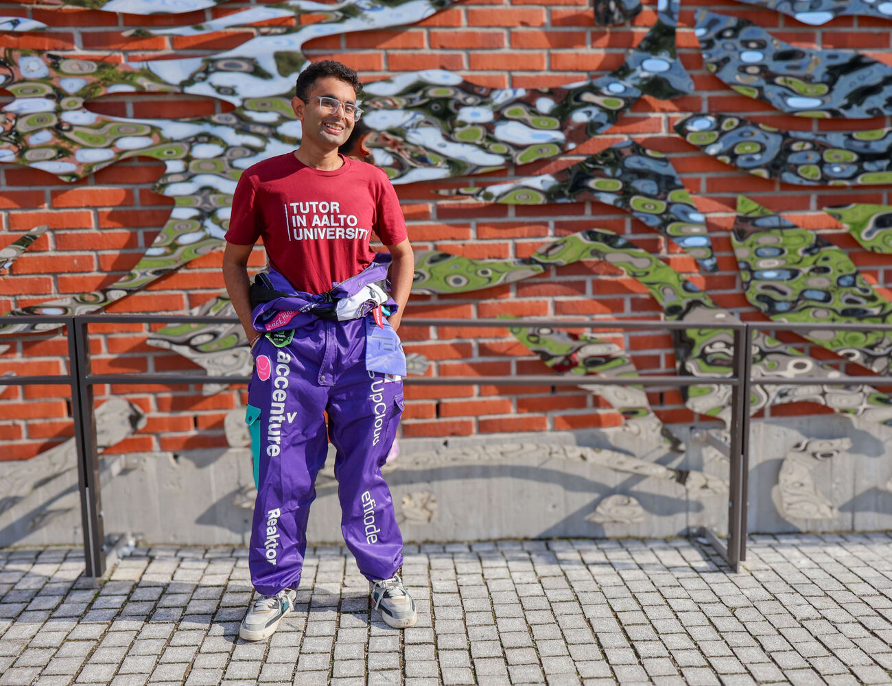 Hitesh Monga wearing Tutor in Aalto University shirt and overalls, standing in front of a brick wall with metal artwork