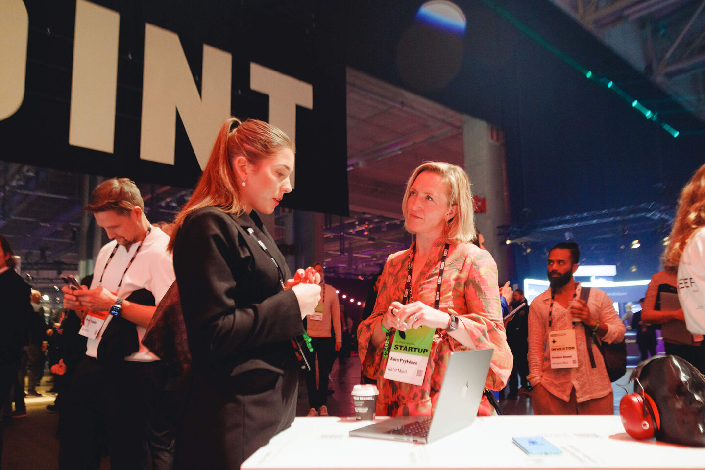 Two women talking at a tech startup event, standing next to a laptop on a table with a coffee cup and headphones nearby.