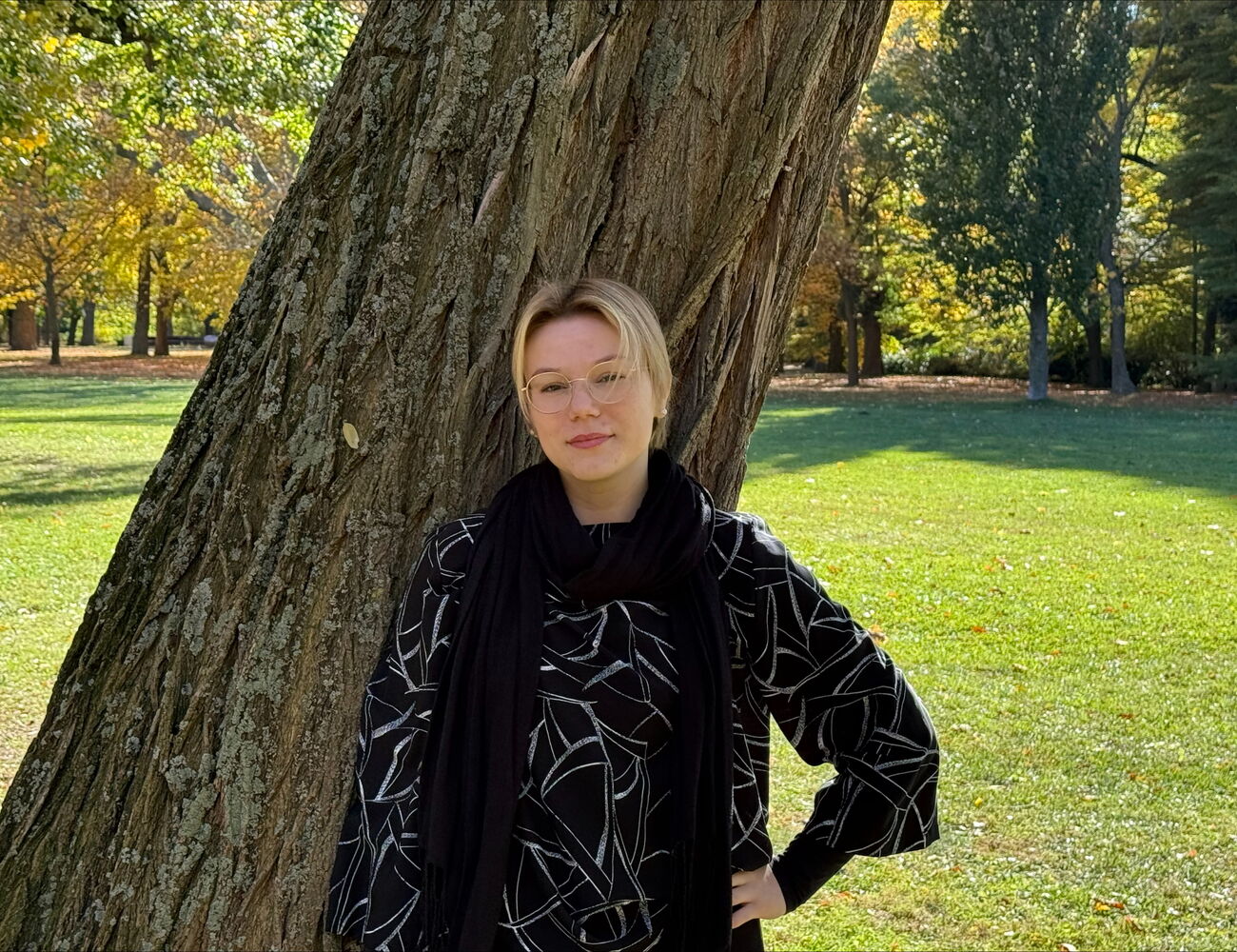 Person with hand on hip leans against large, textured tree trunk in a sunlit park with autumn foliage.