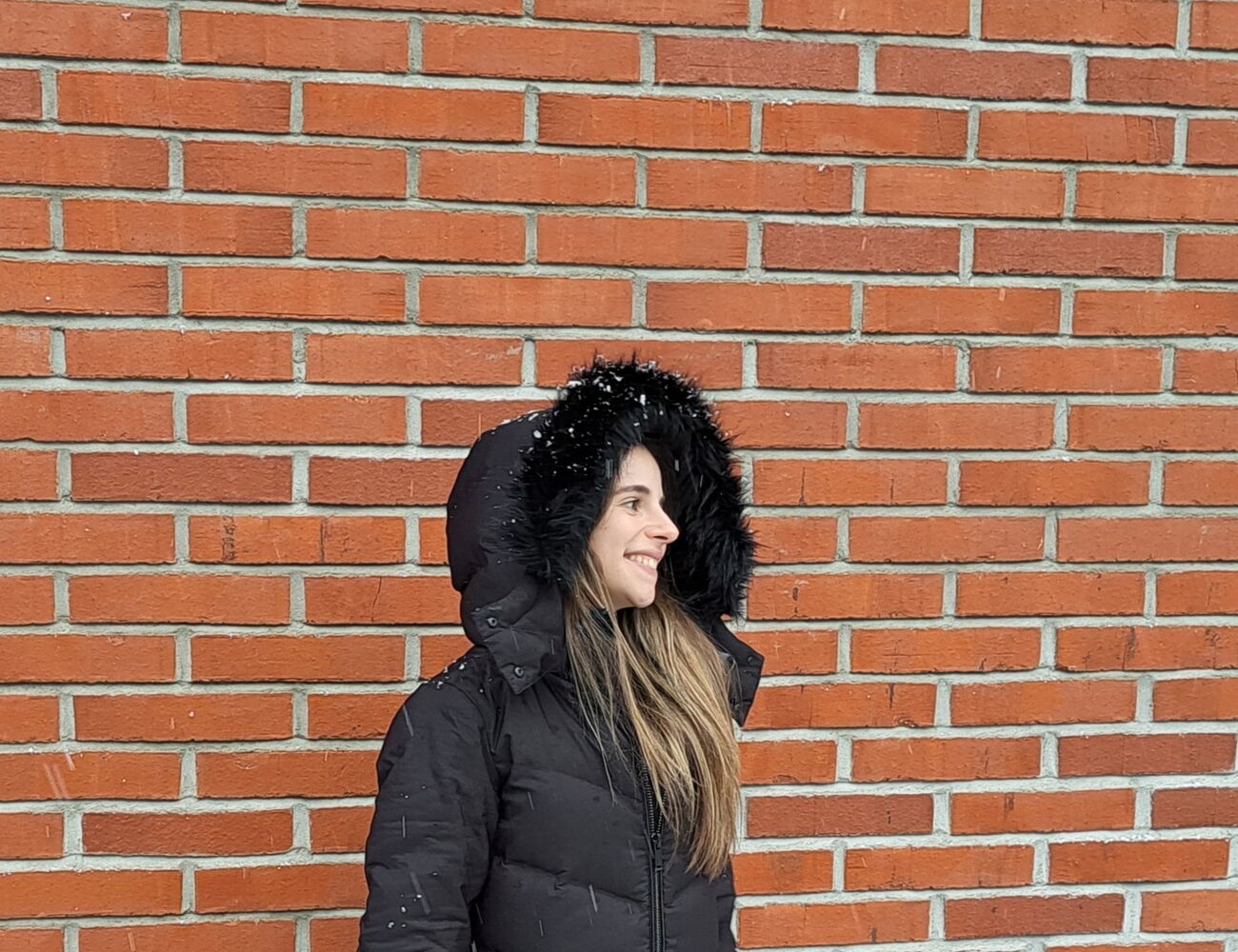 Person wearing a black winter coat with fur hood stands against a red brick wall.