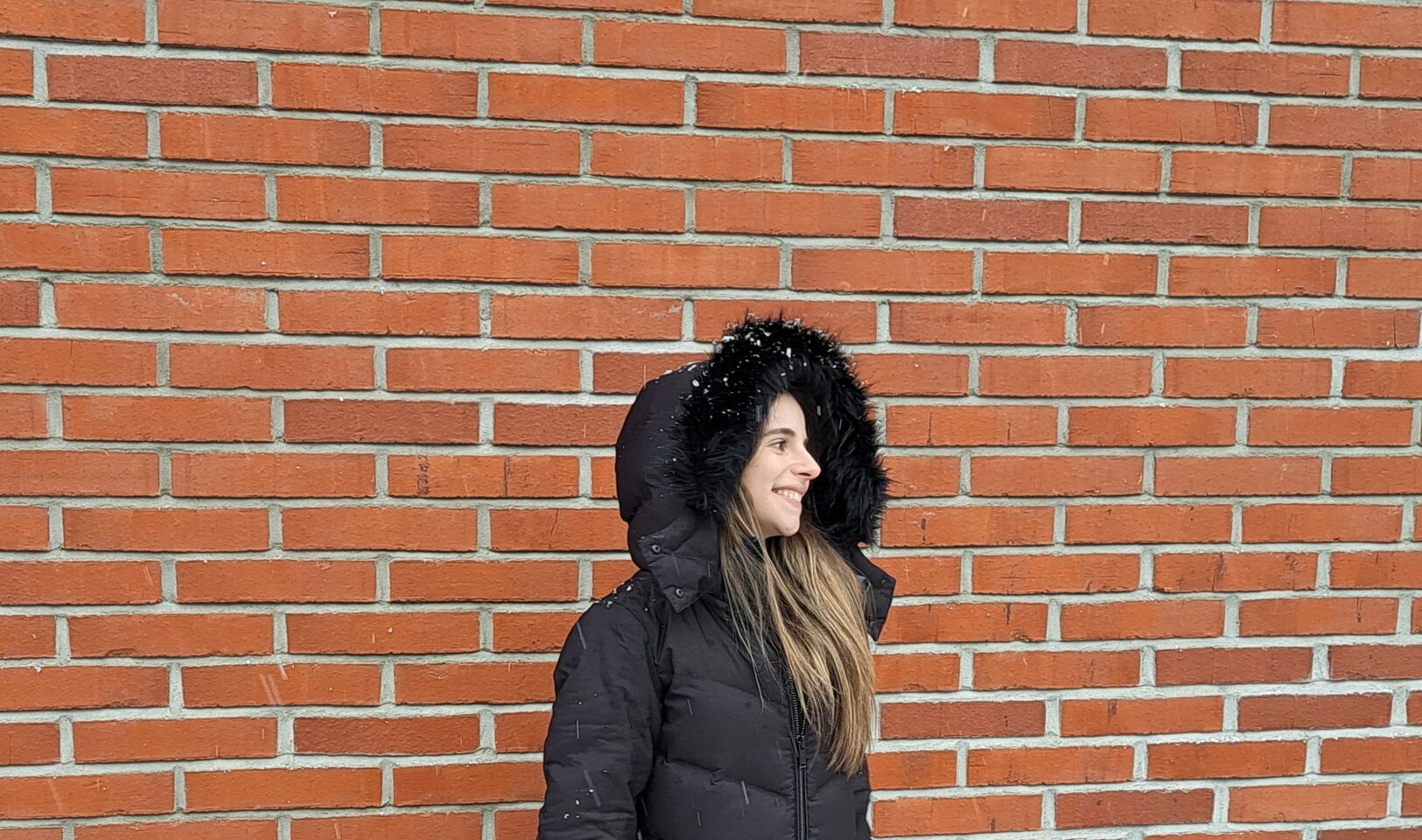 Person wearing a black winter coat with fur hood stands against a red brick wall.