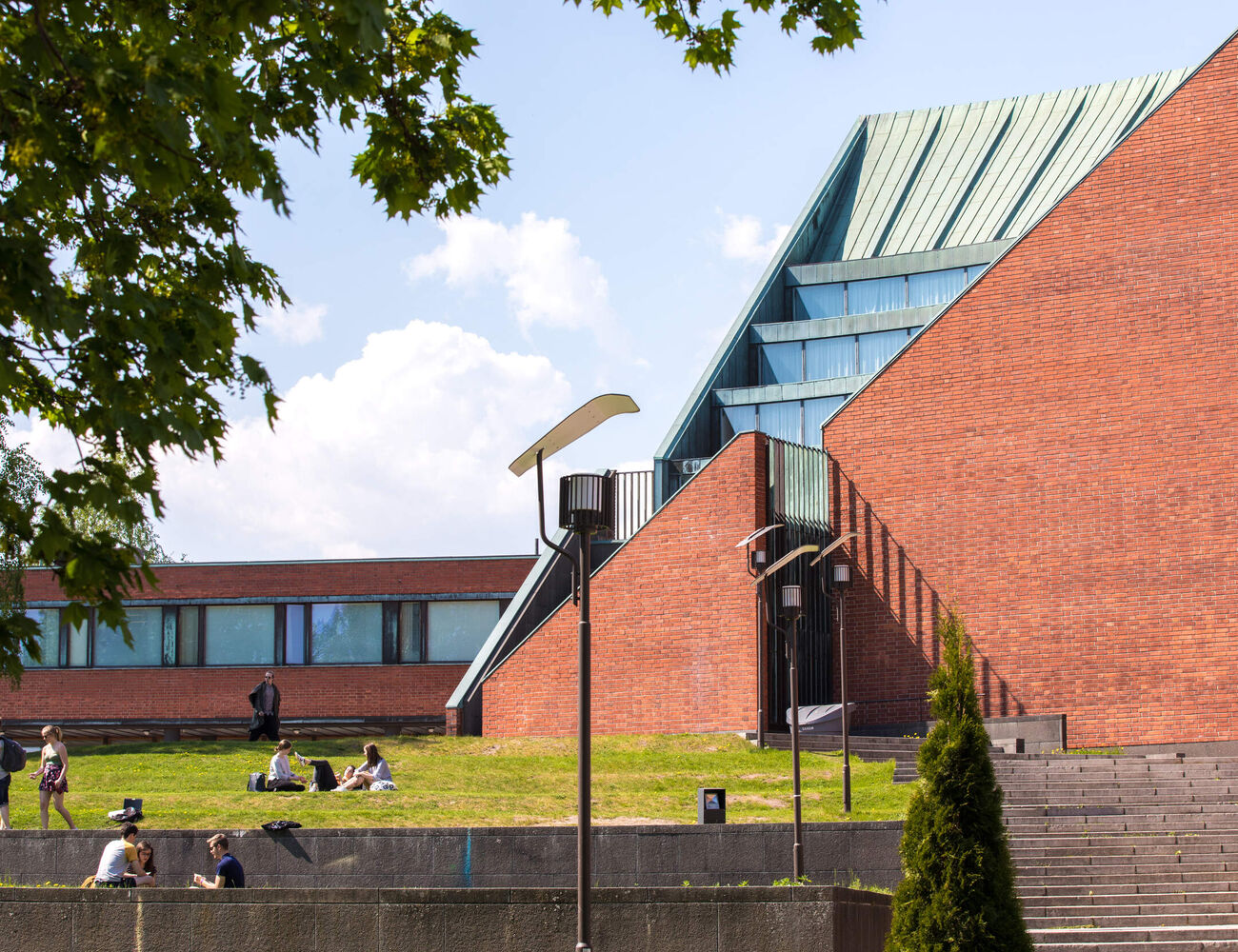 People relax on a grassy area near a modern building with red brick walls and large glass windows under a blue sky.