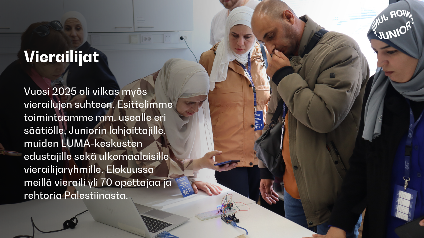 Ryhmä ihmisiä kokoontuneena pöydän ympärille, jossa on elektroniikkakomponentteja ja kannettavia tietokoneita. Teksti vierailijoista vuonna 2025.