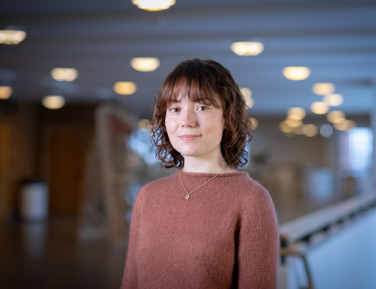 En person med rosa tröja och halsband står inomhus med suddiga ljus i bakgrunden.