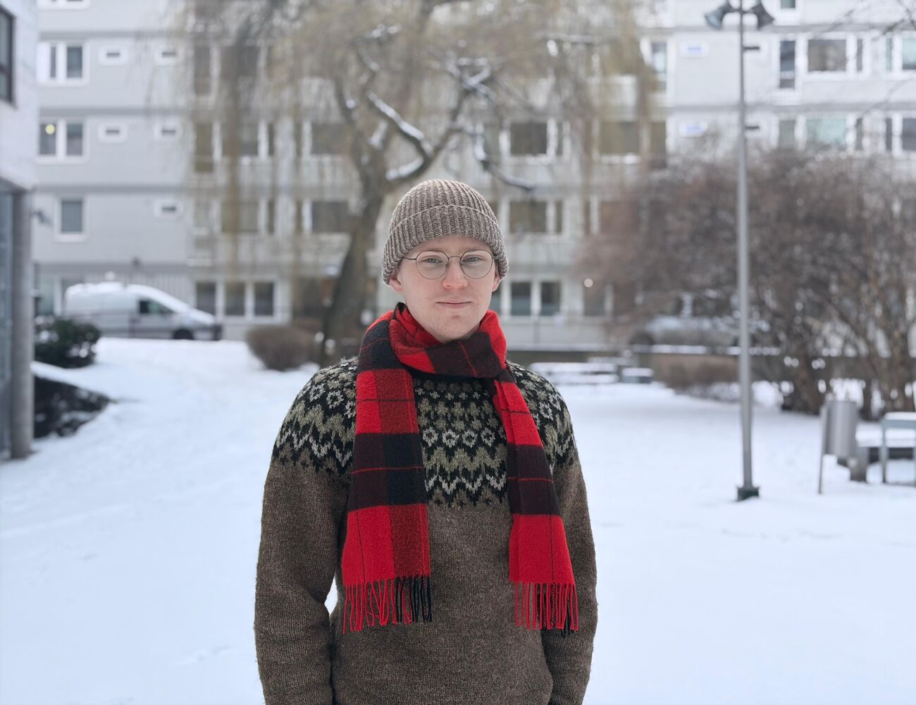 Person i stickad mössa och röd halsduk står på en snöig gård med byggnader och träd i bakgrunden.
