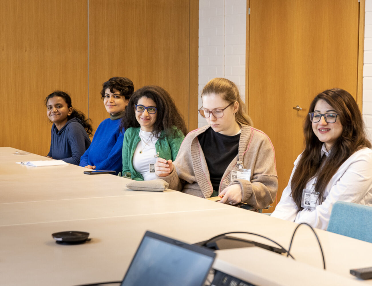 Five people sit at a meeting table in a bright room, listening and talking, with laptops and notes on the table.