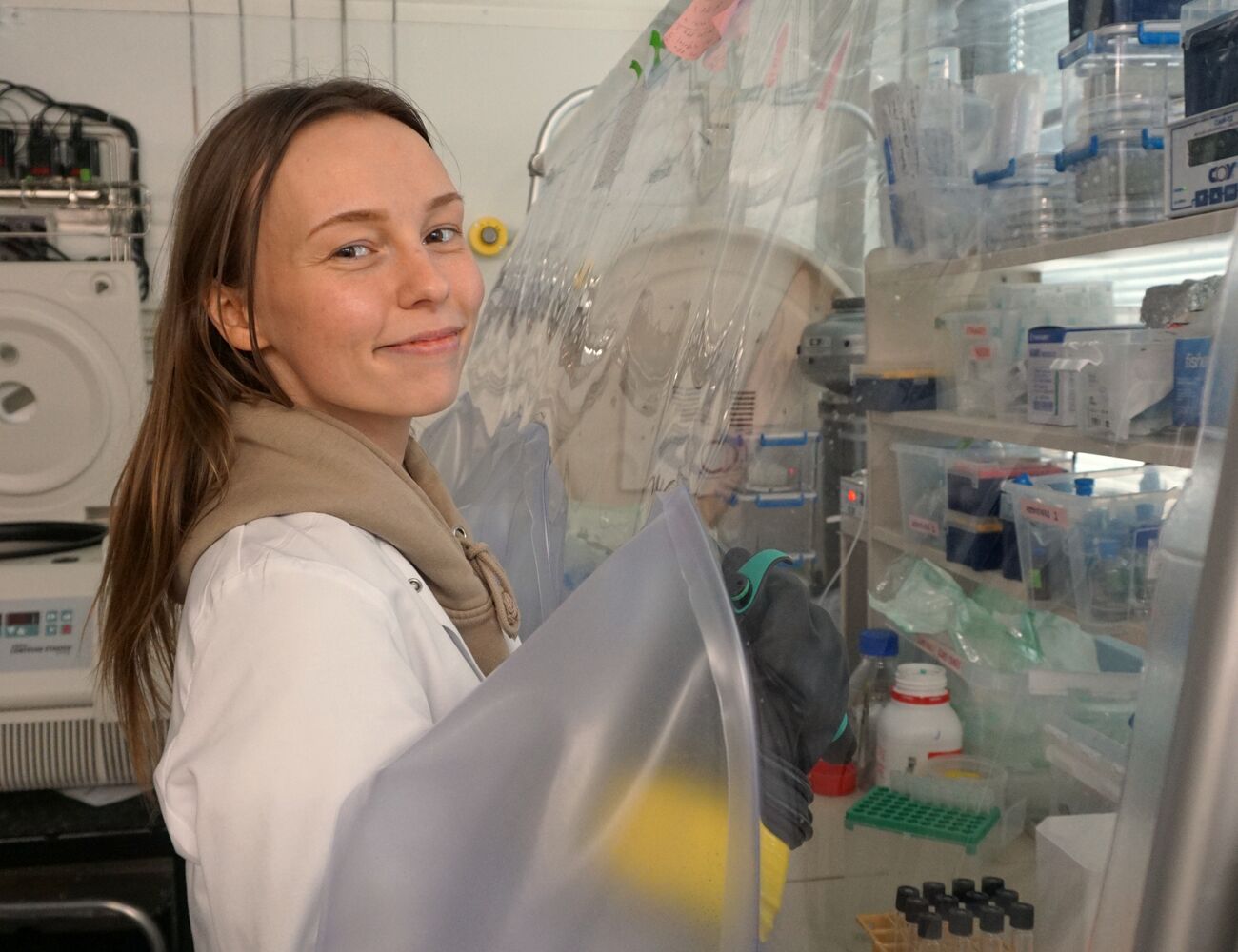 Person i vit rock arbetar vid en handskbox i laboratorium med hyllor fyllda med rör och utrustning