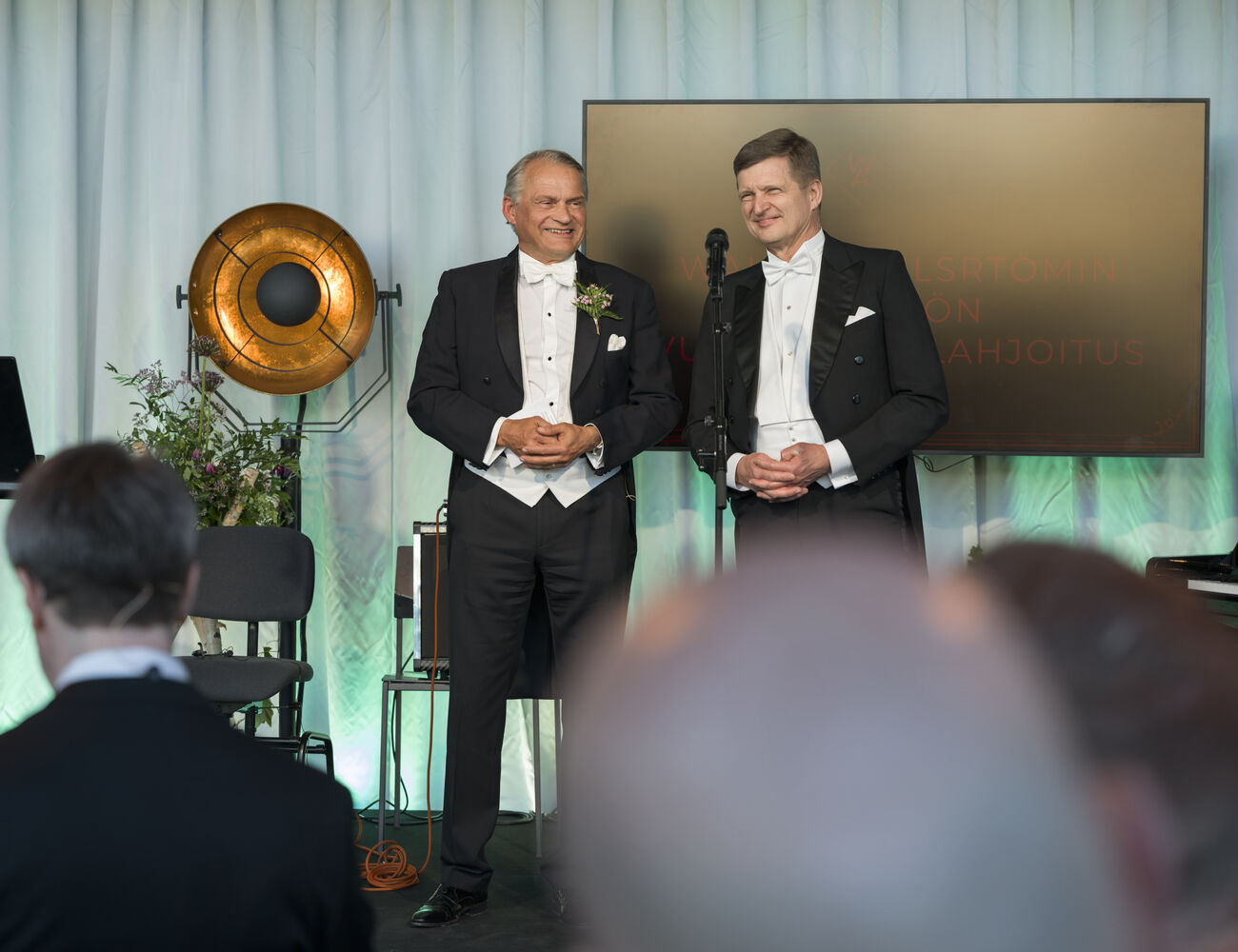 Two men in black tailcoats stand on stage by a microphone, speaking to a seated audience indoors.