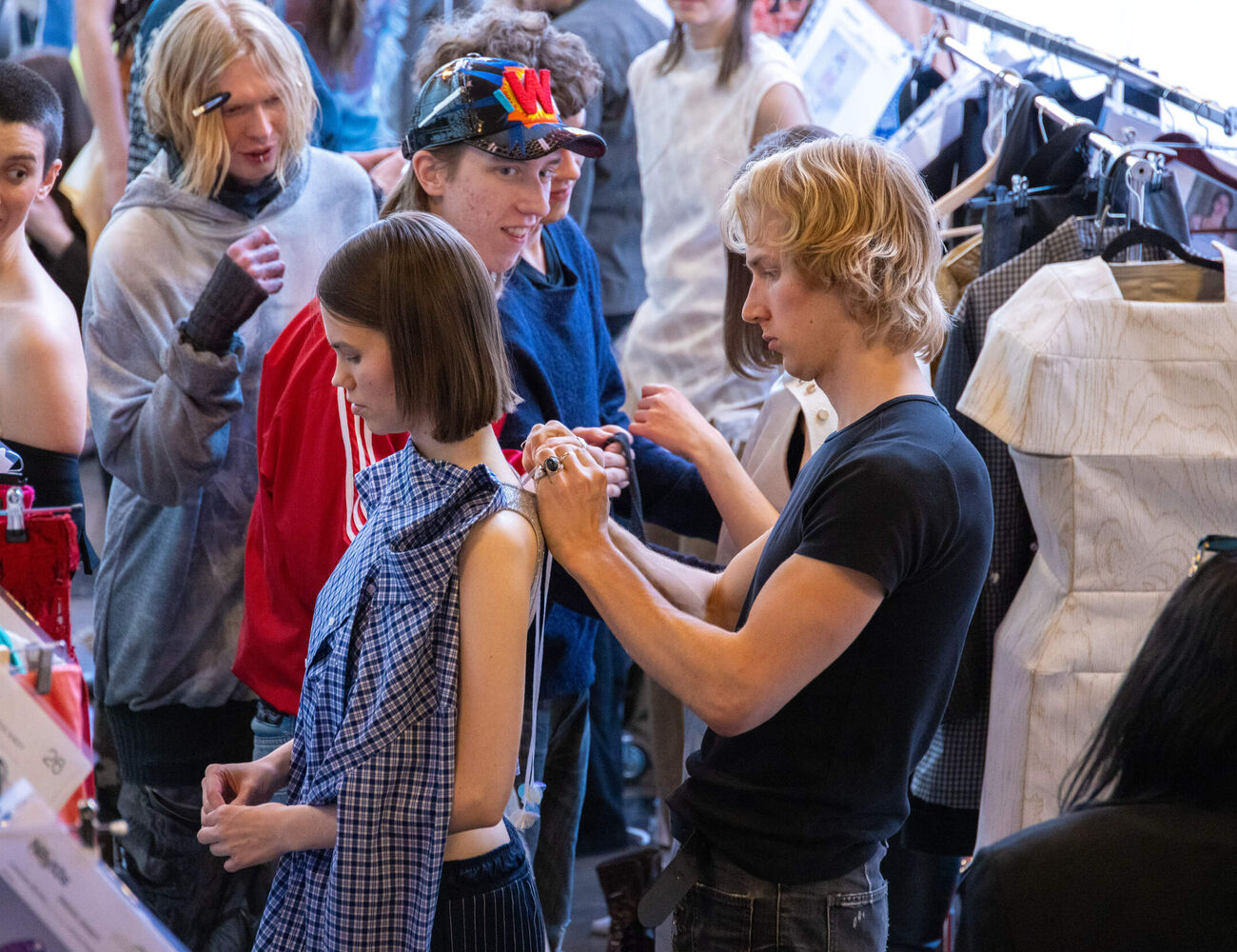Fashion show backstage, stylist adjusting model’s blue check dress amid racks of clothes and people.