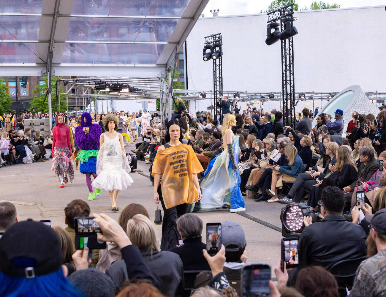 Outdoor fashion show with models in colourful avant-garde outfits walking past a large seated crowd