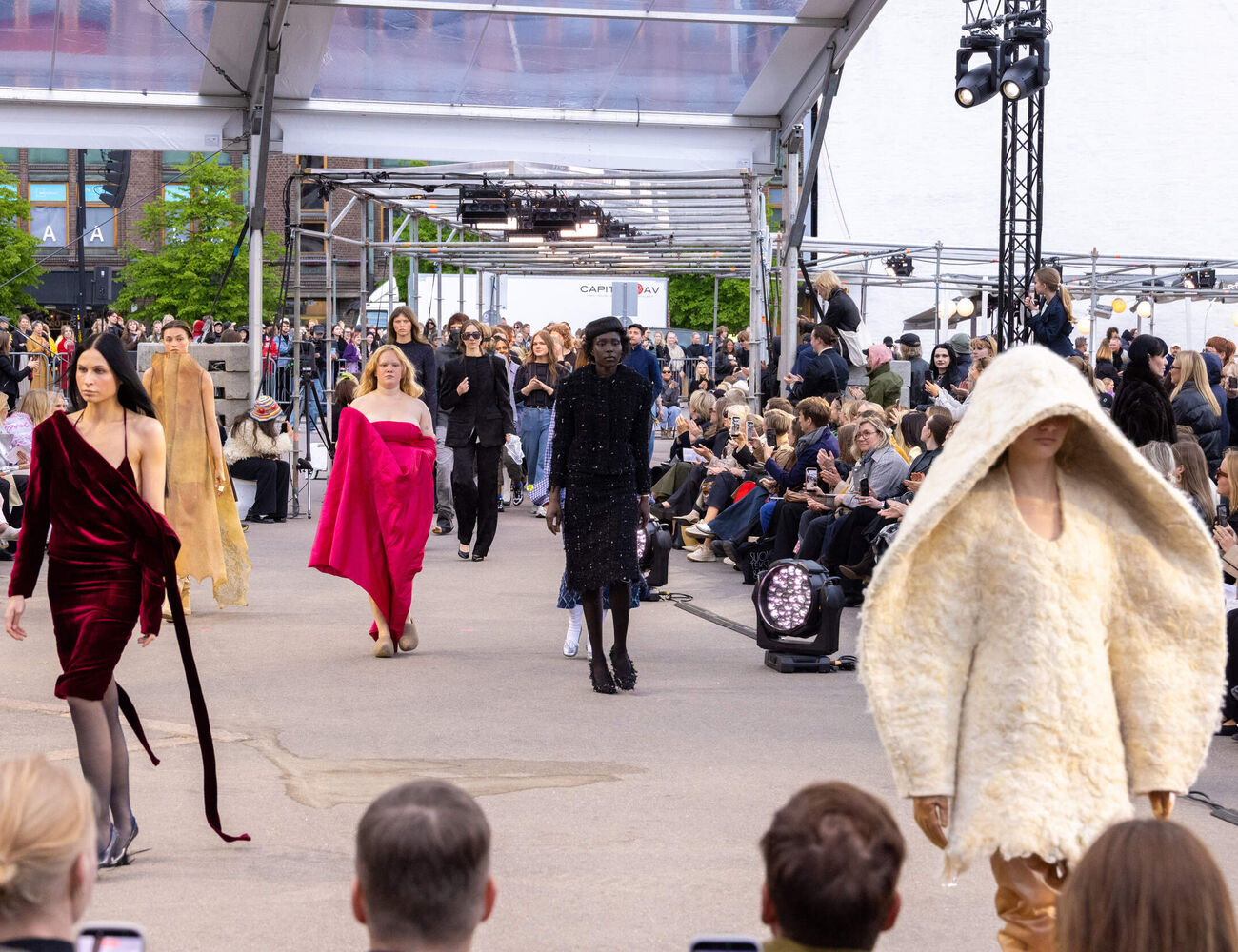 Outdoor fashion show with models in creative outfits walking a catwalk lined by spectators.