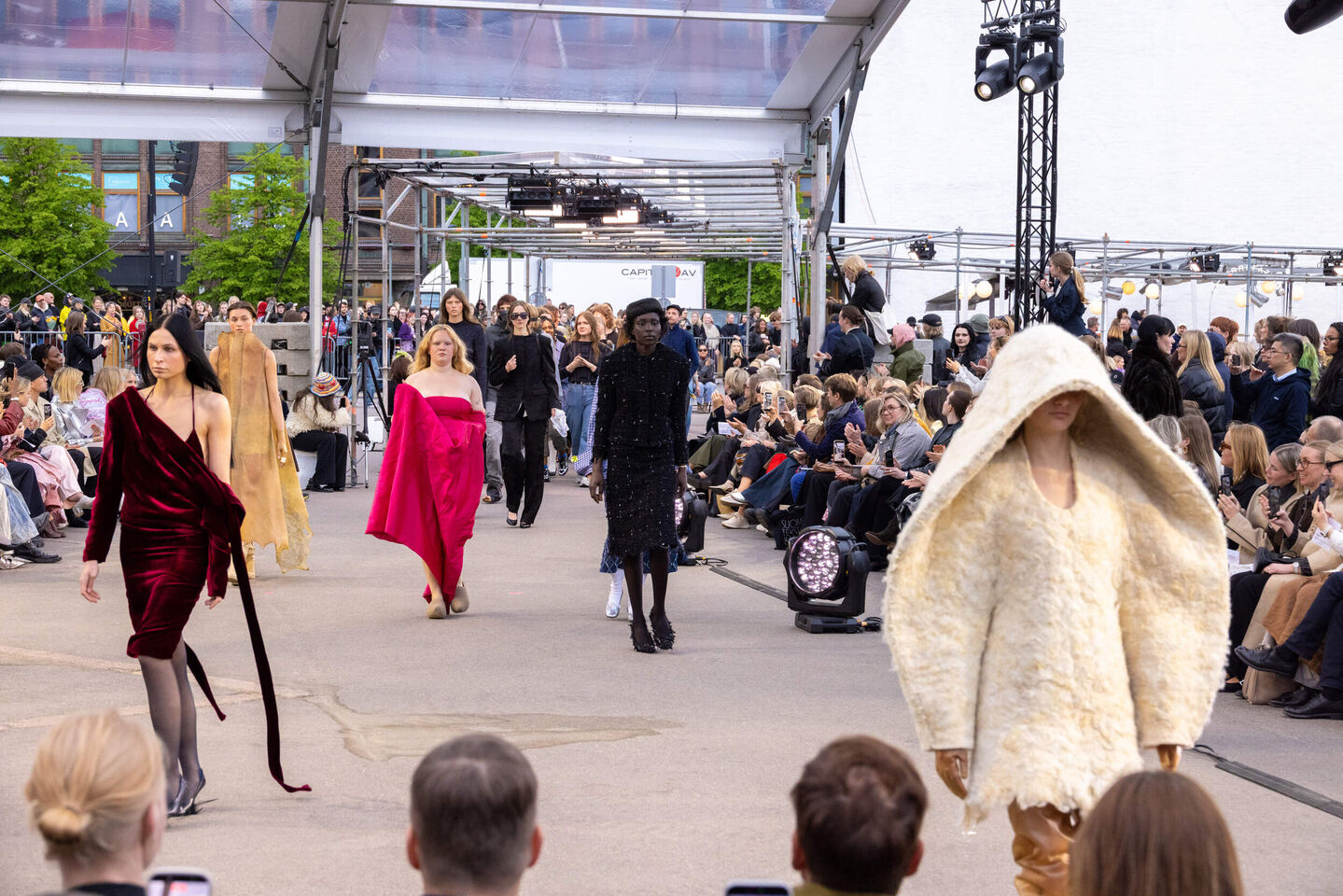 Muotinäytös ulkona, jossa malleja luovissa asuissa kävelee yleisön reunustamalla catwalkilla.