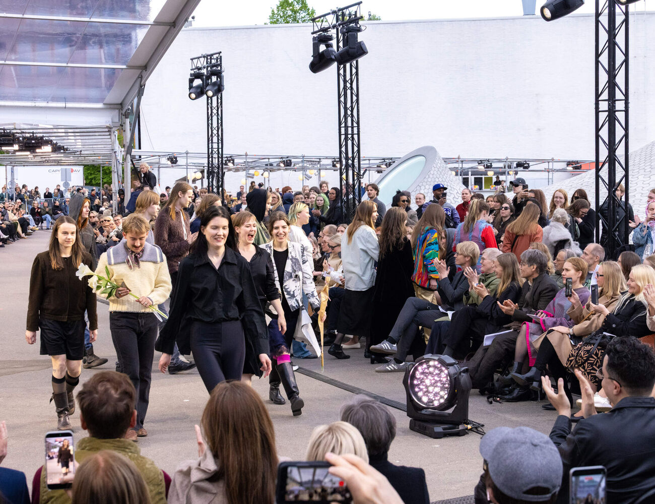 Smiling fashion designers walk on an outdoor runway, audience claps and takes photos.