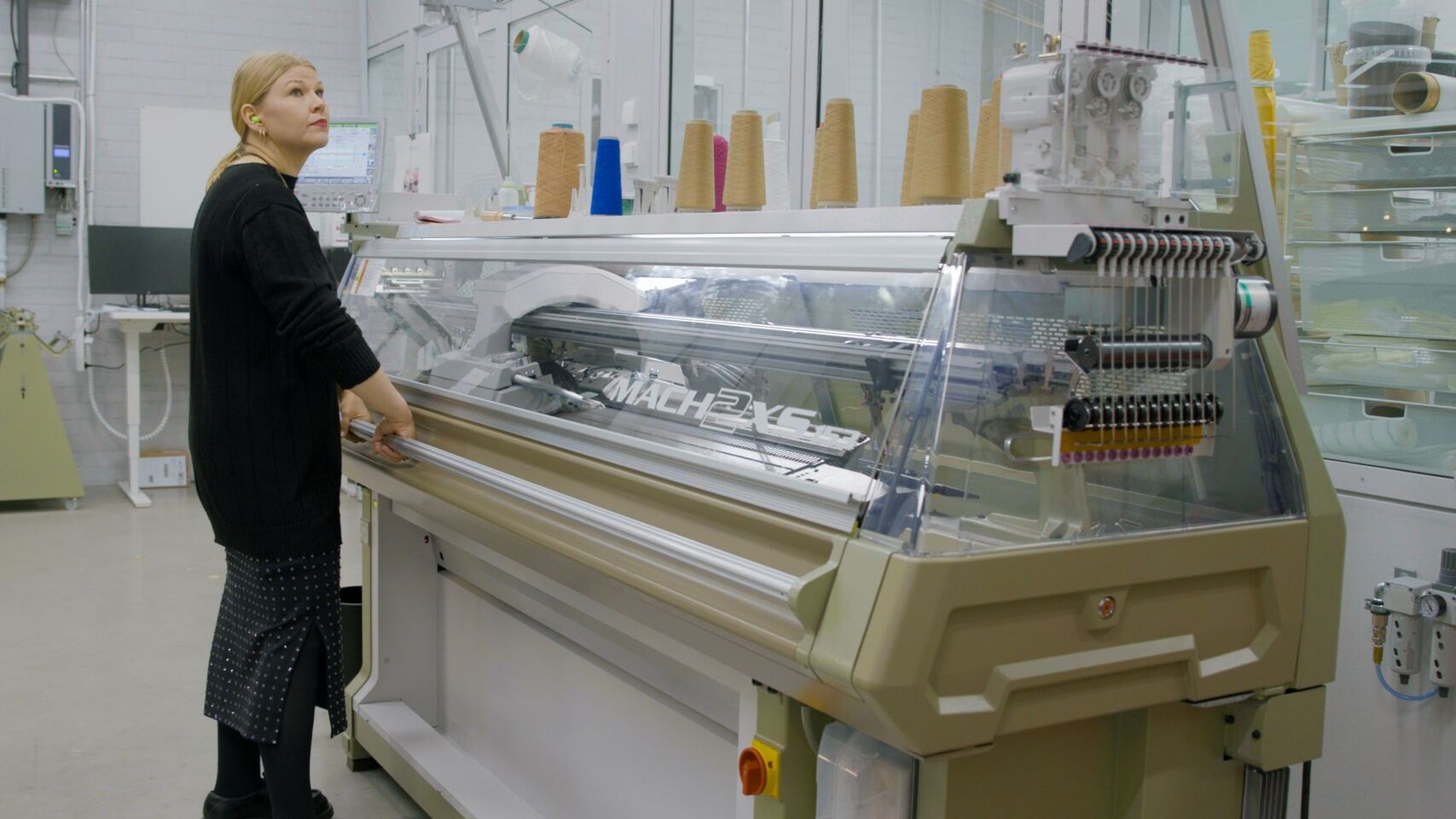 Person in black stands at large beige knitting machine in lab, yarn cones in various colours on top.