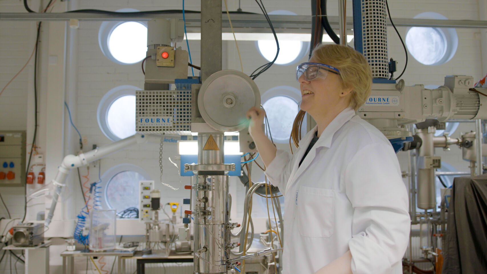 Person in white lab coat operating tall industrial machine in bright laboratory with round windows
